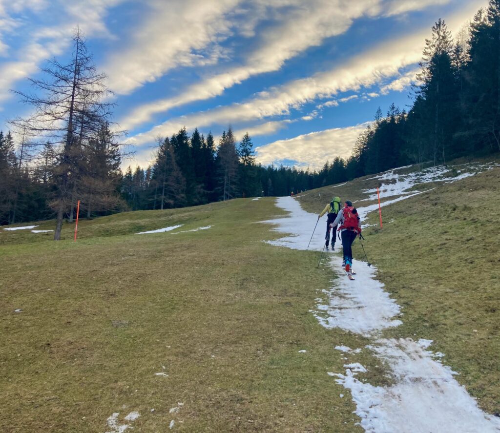 Schmaler Streifen Schnee auf grüner Wiese mit zwei Skitourengängern.